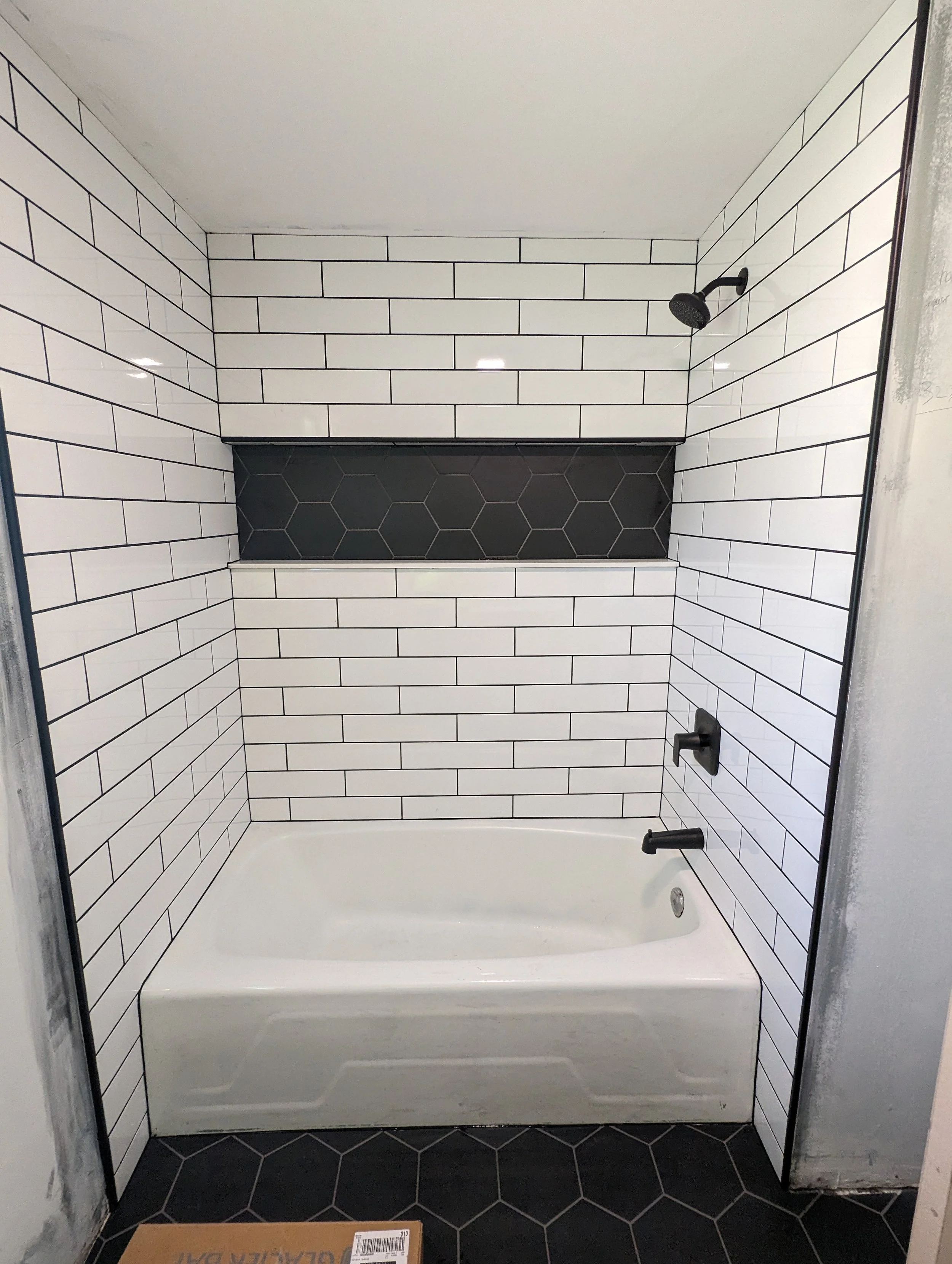 A bathroom shower with white subway tile walls, a black hexagonal tile shelf, a black showerhead, and black fixtures. A black handrail is installed on the right wall and a cardboard box is on the black hex tile floor.