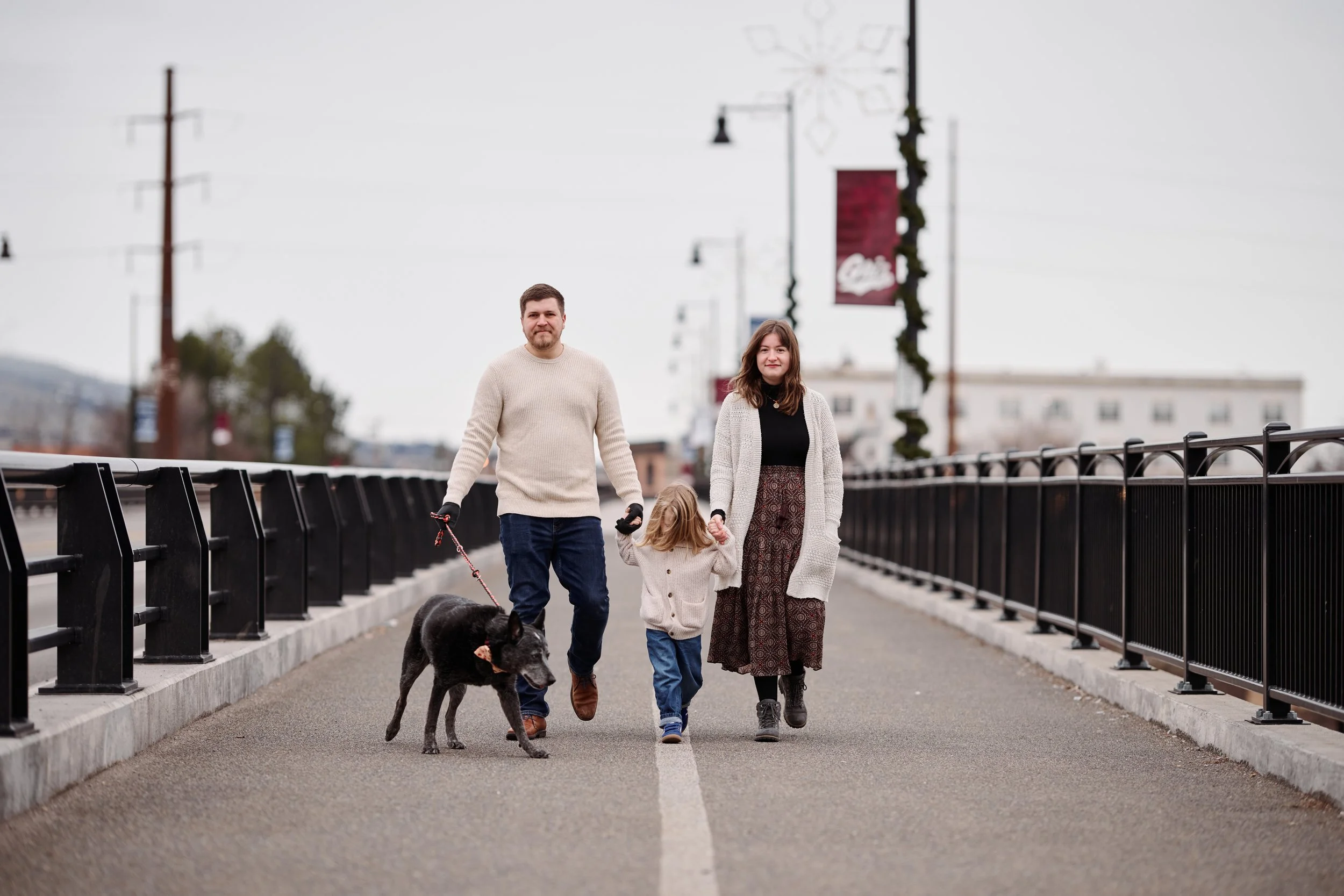 A family of four walking along a bridge sidewalk with their dog. The father is holding the dog's leash, and the mother and daughter are walking hand-in-hand.
