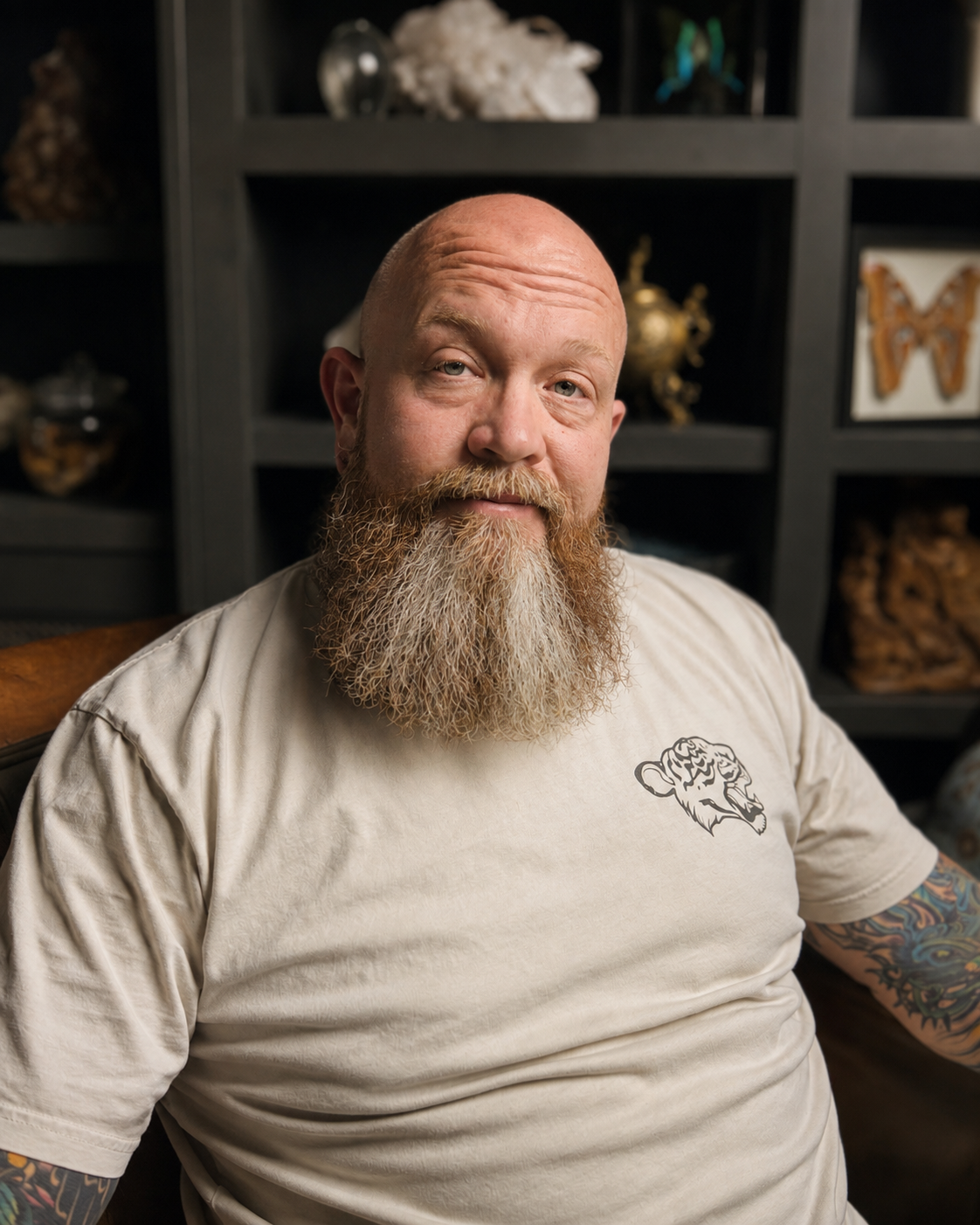 A bald man with a long, full beard and tattoos on his arms, wearing a beige T-shirt with a lion tattoo design, sitting in front of a dark shelf filled with decorative objects and artwork.