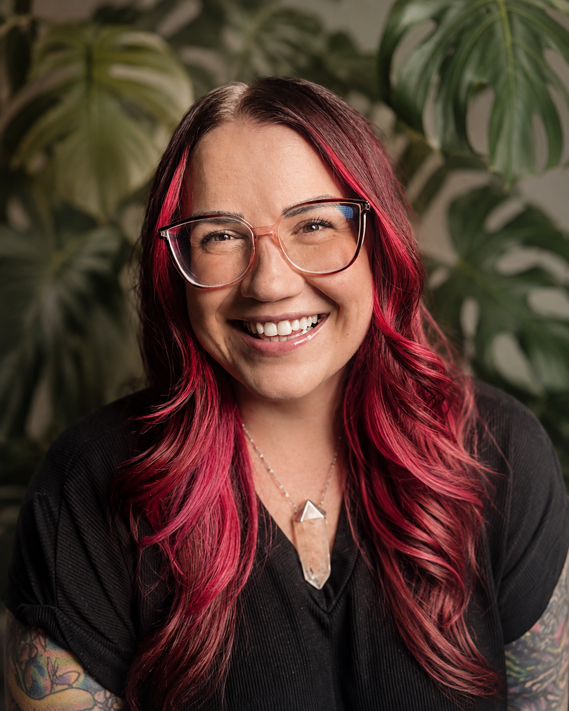 A woman with pink dyed hair, wearing glasses, smiling in front of green leafy plants.