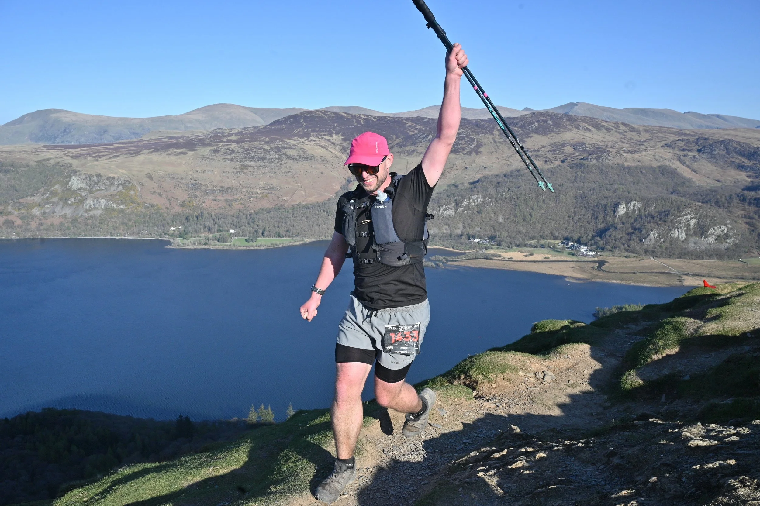 A man in athletic gear, wearing a pink cap and sunglasses, smiling and raising his arm with trekking poles during a trail run near a lake with mountains in the background.