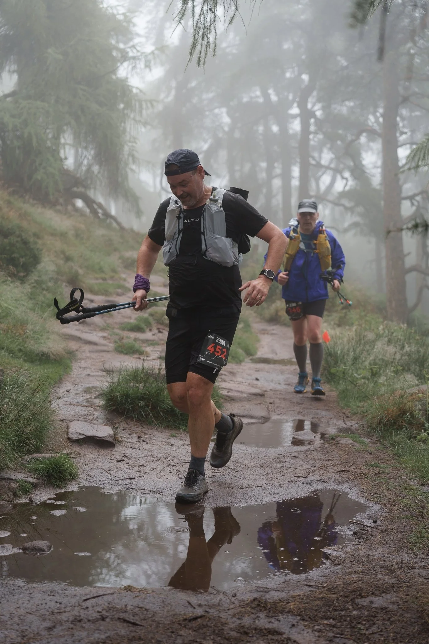 Two men trail running on a muddy, wet forest trail surrounded by trees and fog, with puddles on the ground reflecting their images.