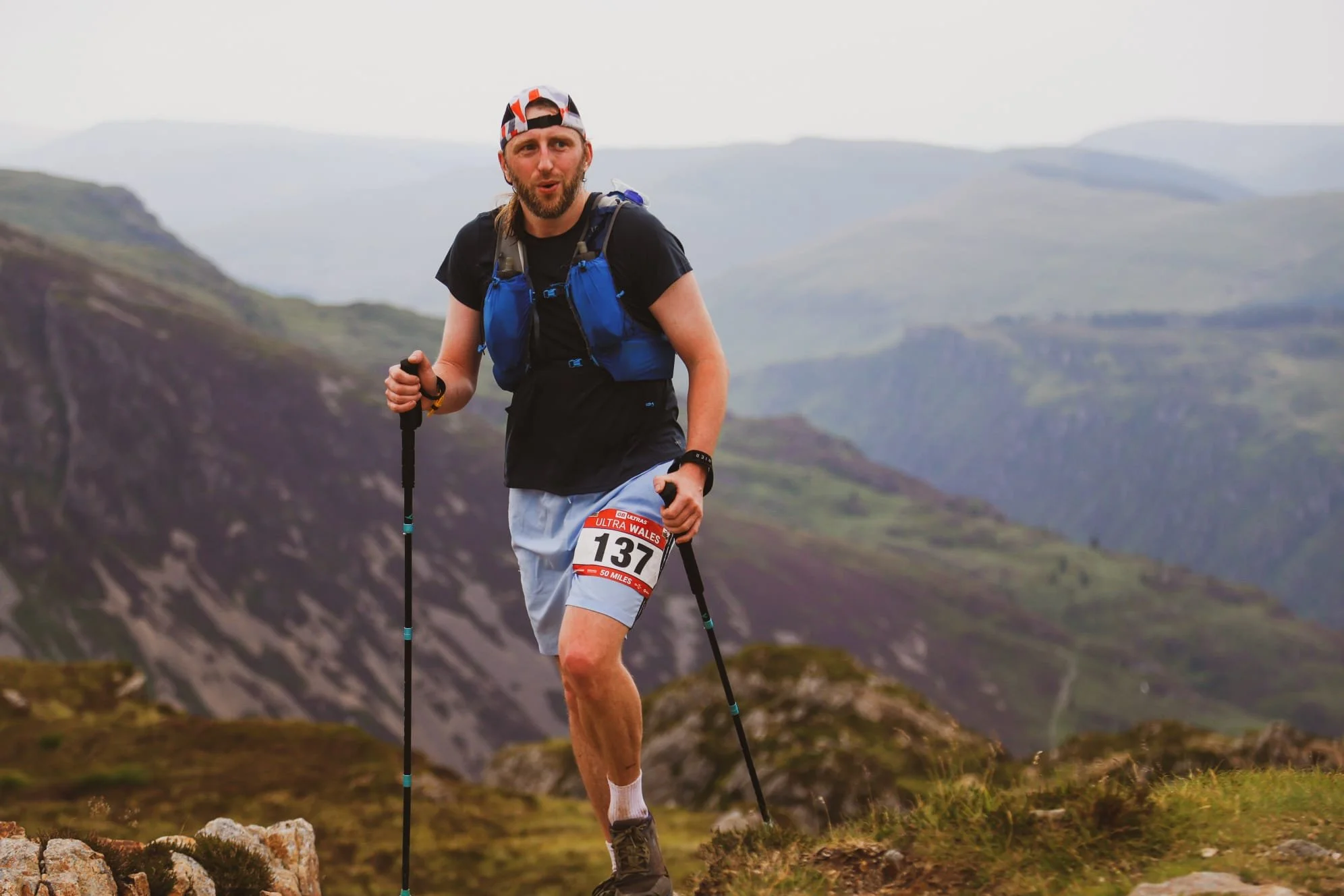 Man trail running in a mountainous landscape, wearing a black shirt, gray shorts with race bib number 137, a blue hydration vest, a white and red cap, and using trekking poles.