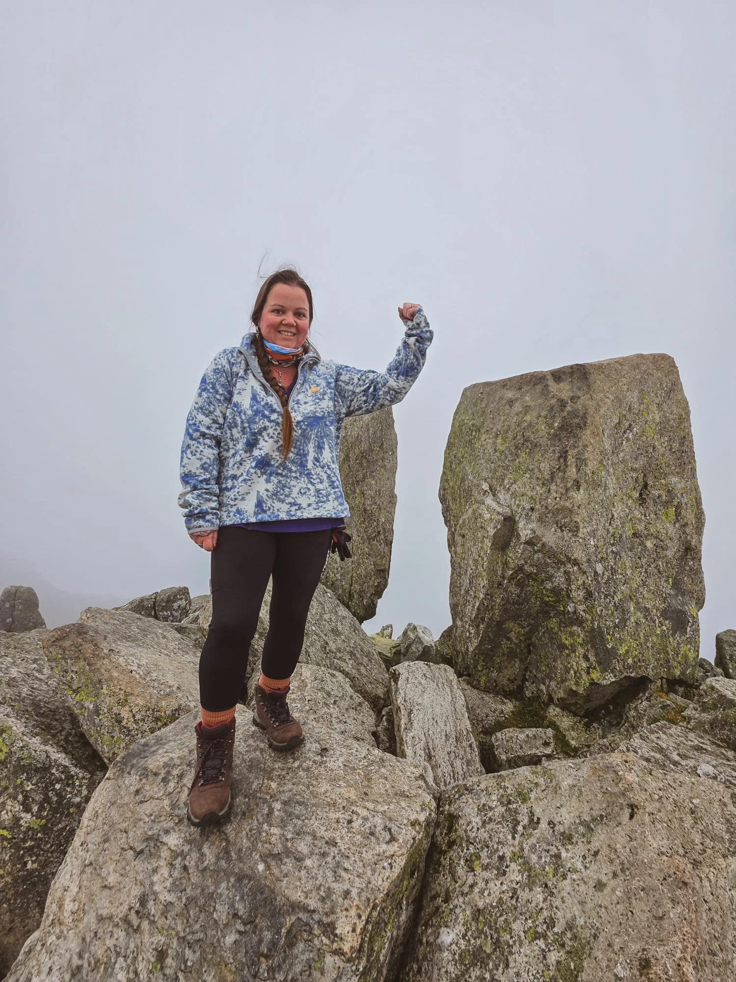 Woman standing on large rocks at mountain summit, pointing at a stone marker, wearing outdoor gear and smiling.