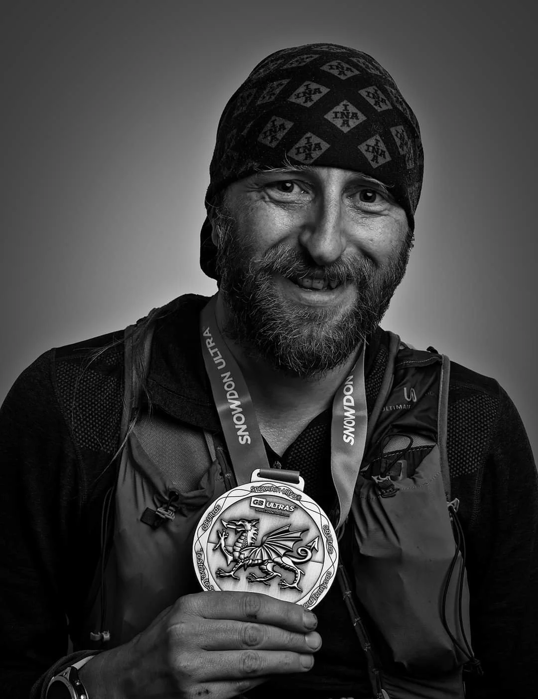 Man with a beard and mustache smiling, wearing a head covering and athletic gear, displaying a medal with a dragon design.