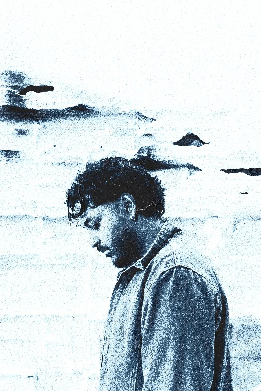 A young man with wet, curly hair and a denim jacket standing outdoors against a wall with rocks in the background.