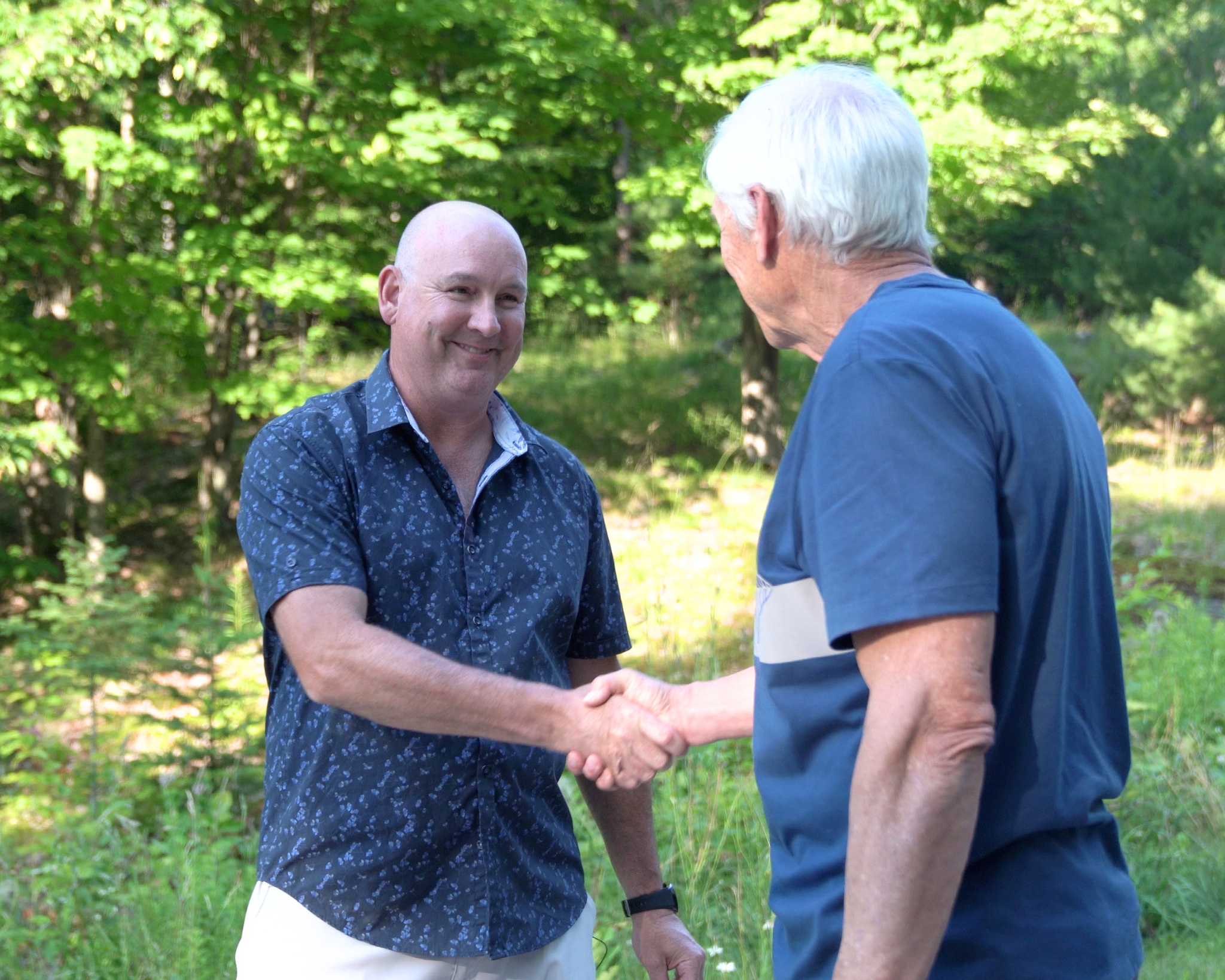 Two men shaking hands outdoors in a green, leafy park, smiling at each other.