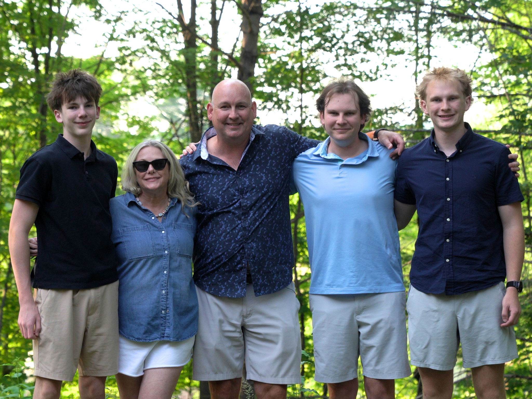 The Caswell Family standing outdoors on their deck, smiling at the camera with their hands all wrapped around each other. They are dressed in casual summer clothing. From left to right is Matthew, Pam, Dan's Wife, Dan,  William, and Nolan