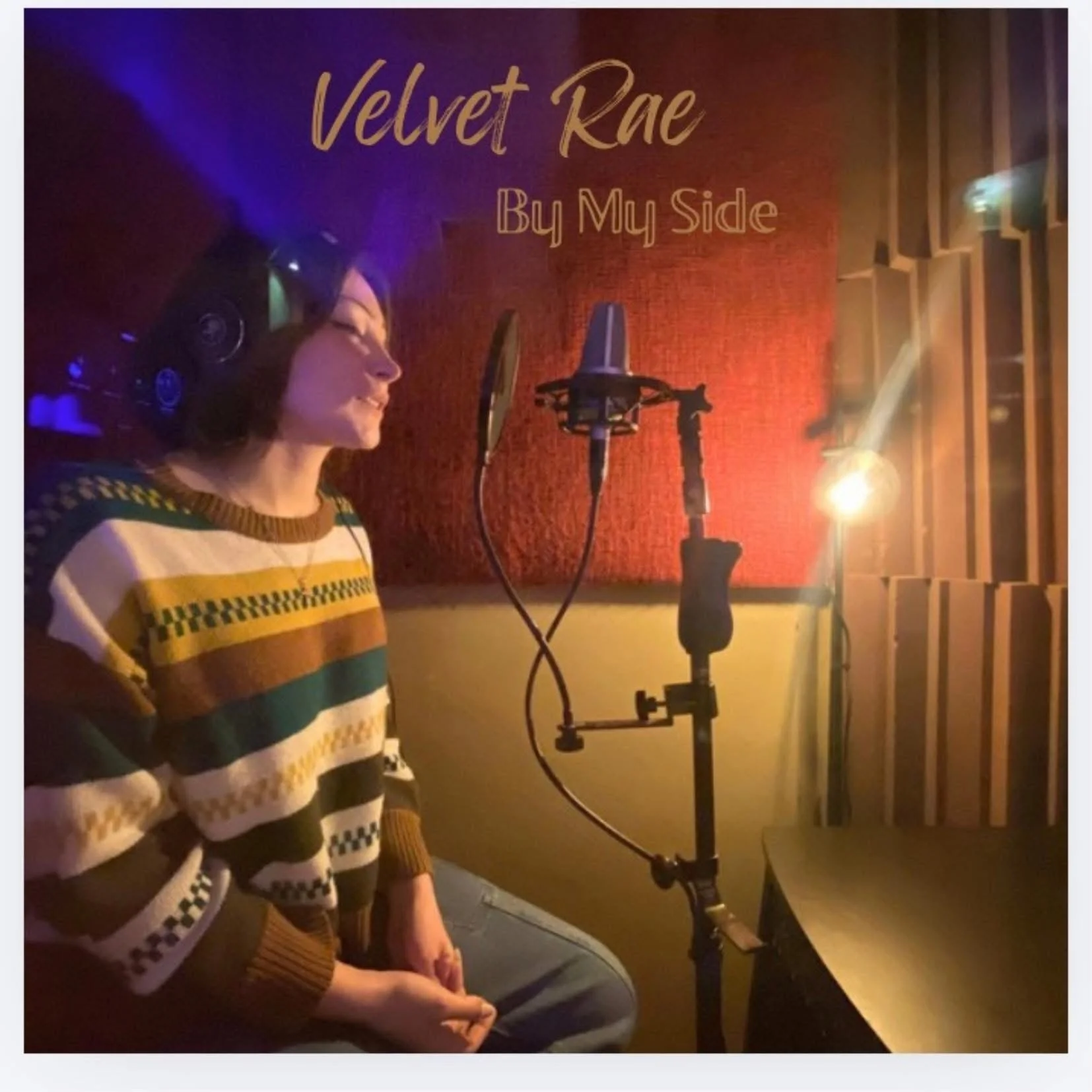A woman in a striped sweater sitting in a recording studio with a microphone and headphones, with the sign 'Velvet Rae By My Side' on the red wall behind her.