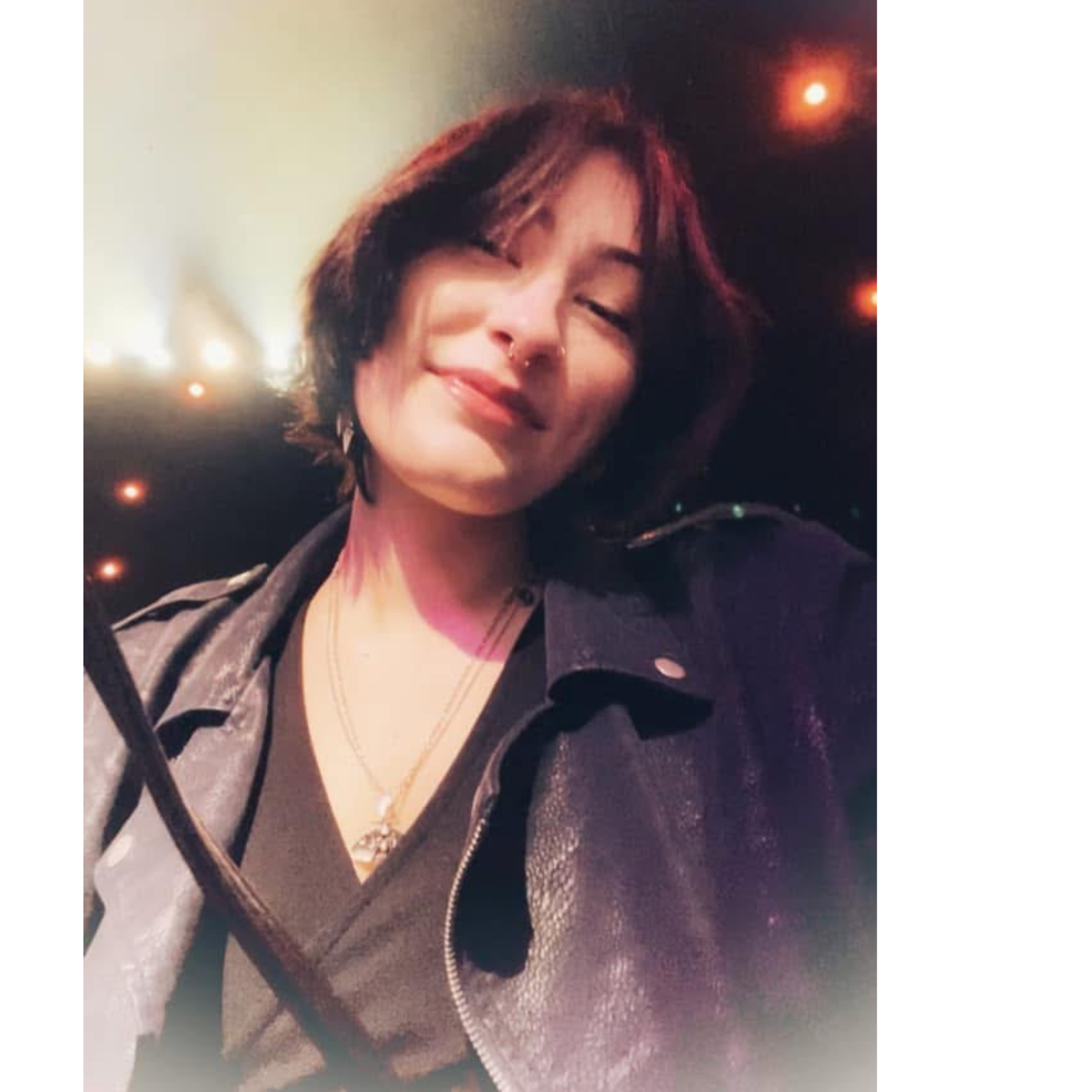A woman with short hair smiling, wearing a dark jacket with a textured tan collar, gold necklaces, and a nose ring, with a blurred background of string lights.
