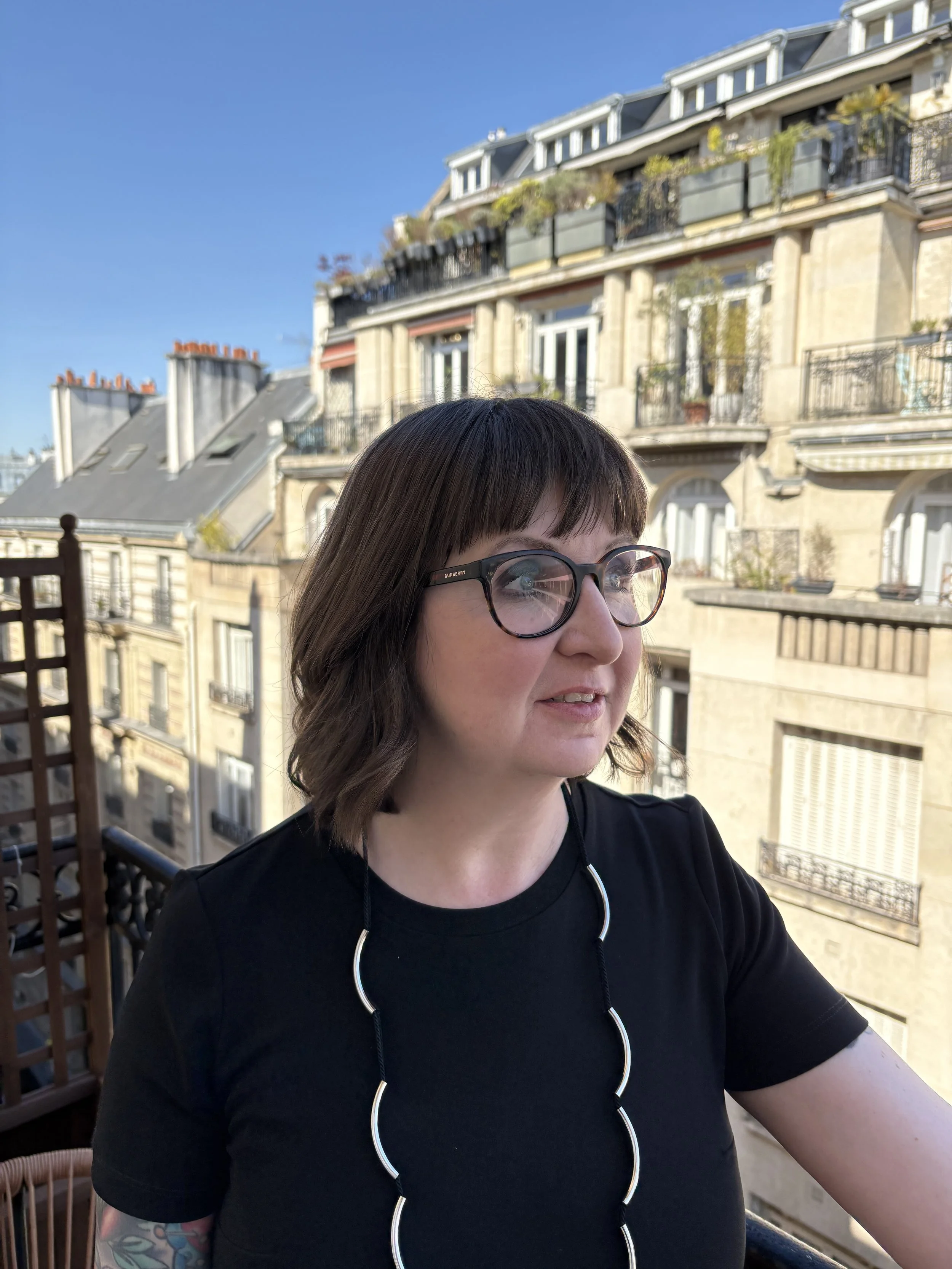 A woman with short brown hair wearing glasses and a black t-shirt, taking a selfie on a balcony with European-style buildings in the background under a clear blue sky.
