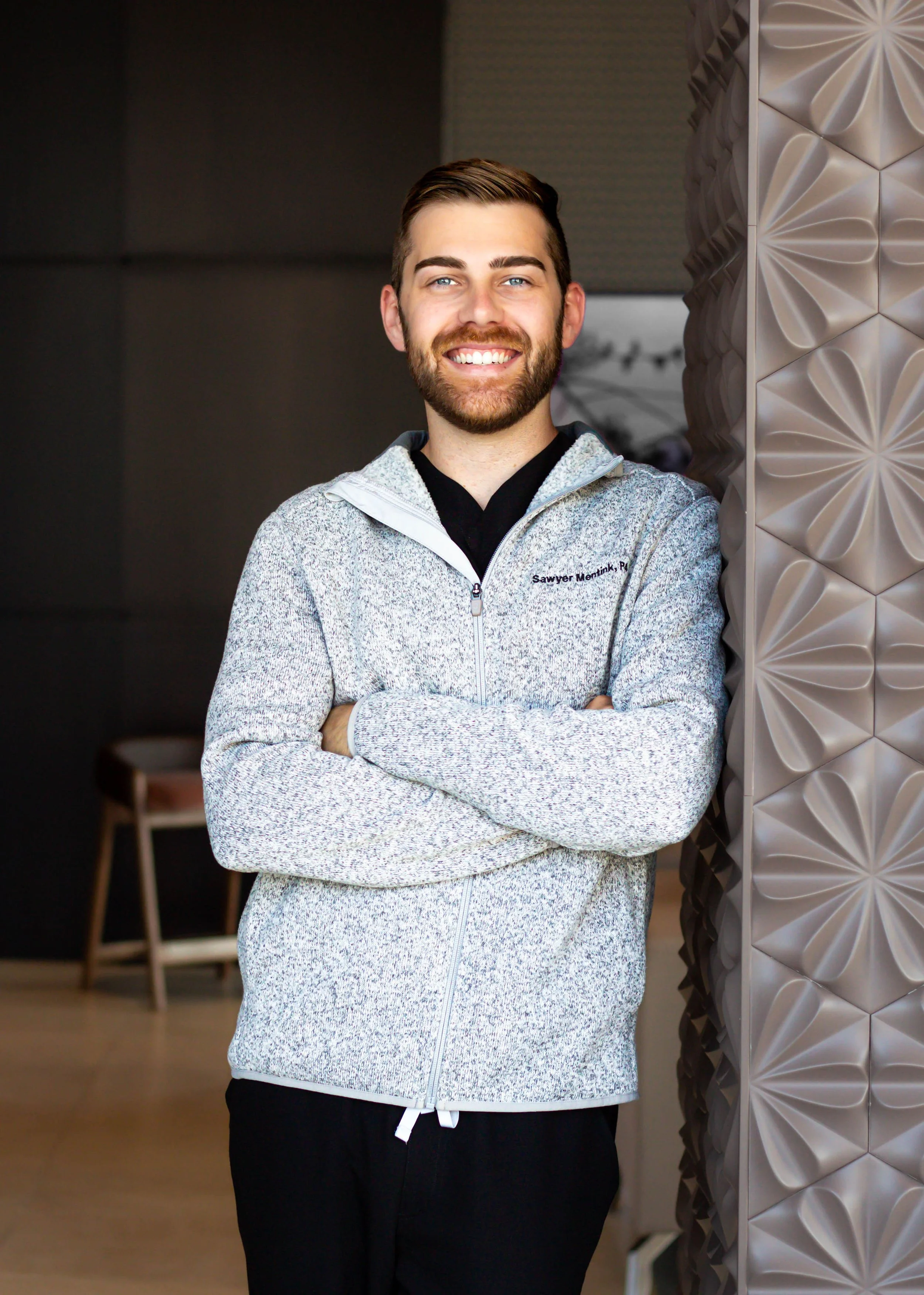A smiling young man with a beard and blue eyes, standing with arms crossed, wearing a gray zip-up jacket and black pants, leaning against a textured wall in an indoor setting.