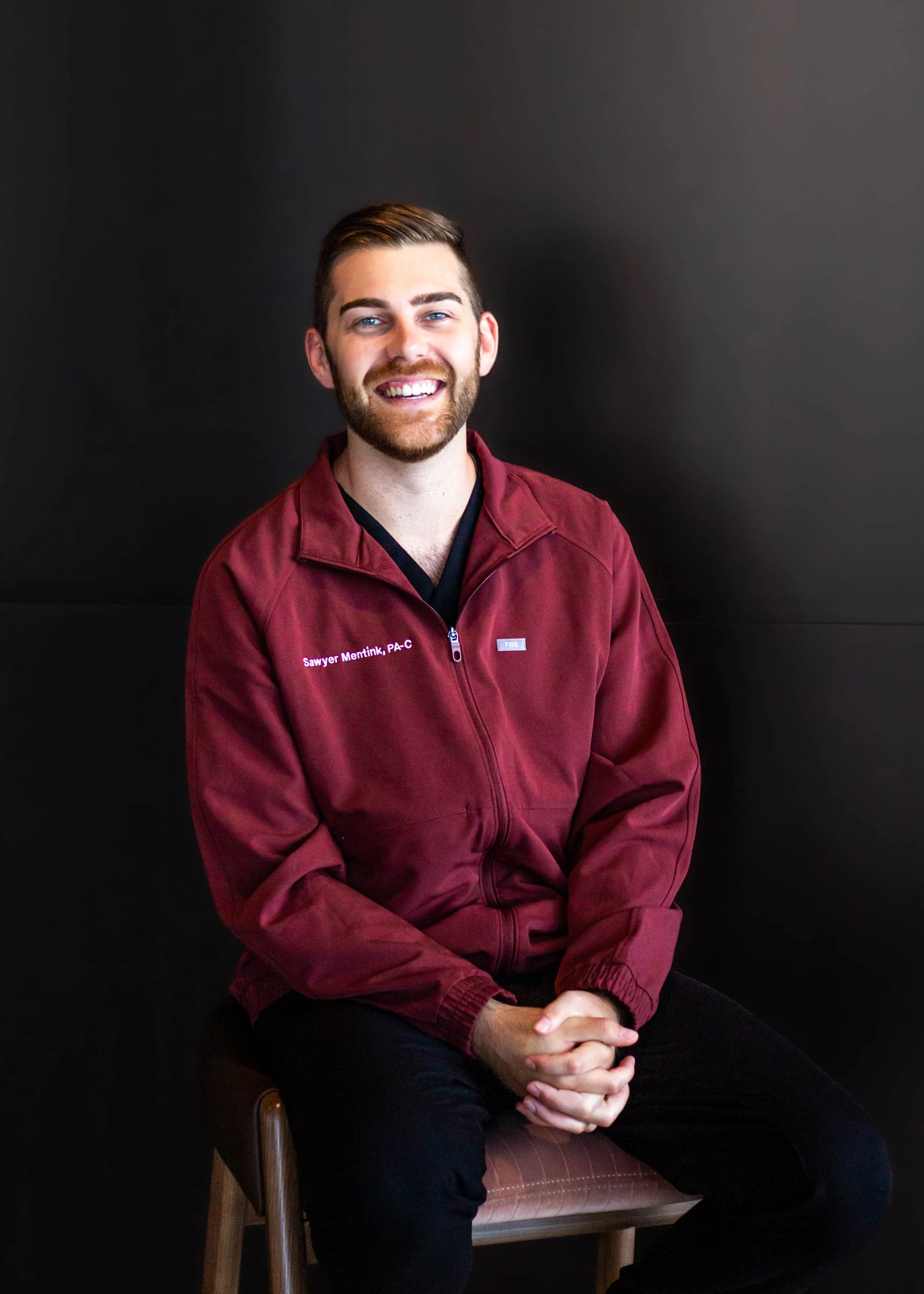 A young man with brown hair and a beard, smiling, sitting on a wooden chair against a dark background. He is wearing a maroon jacket with text on the left side that reads "Sawyer Mentink, PA-C." His hands are clasped together in his lap.