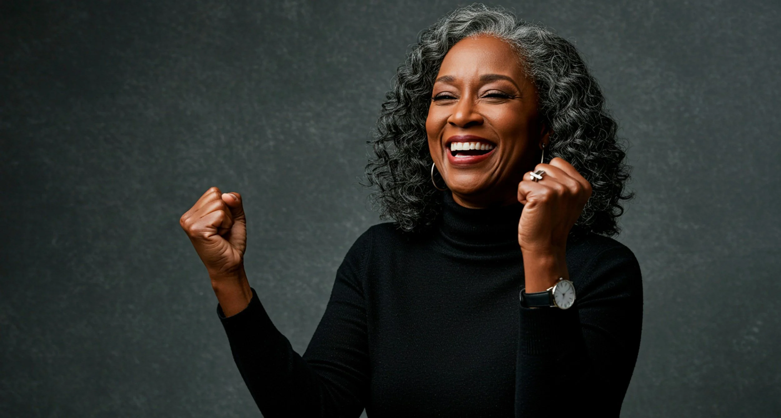 A joyful middle-aged African American woman in her 50's with curly gray hair, wearing a black turtleneck, sporting a watch and earrings, smiling and flexing her arms in a victory pose against a textured dark background.