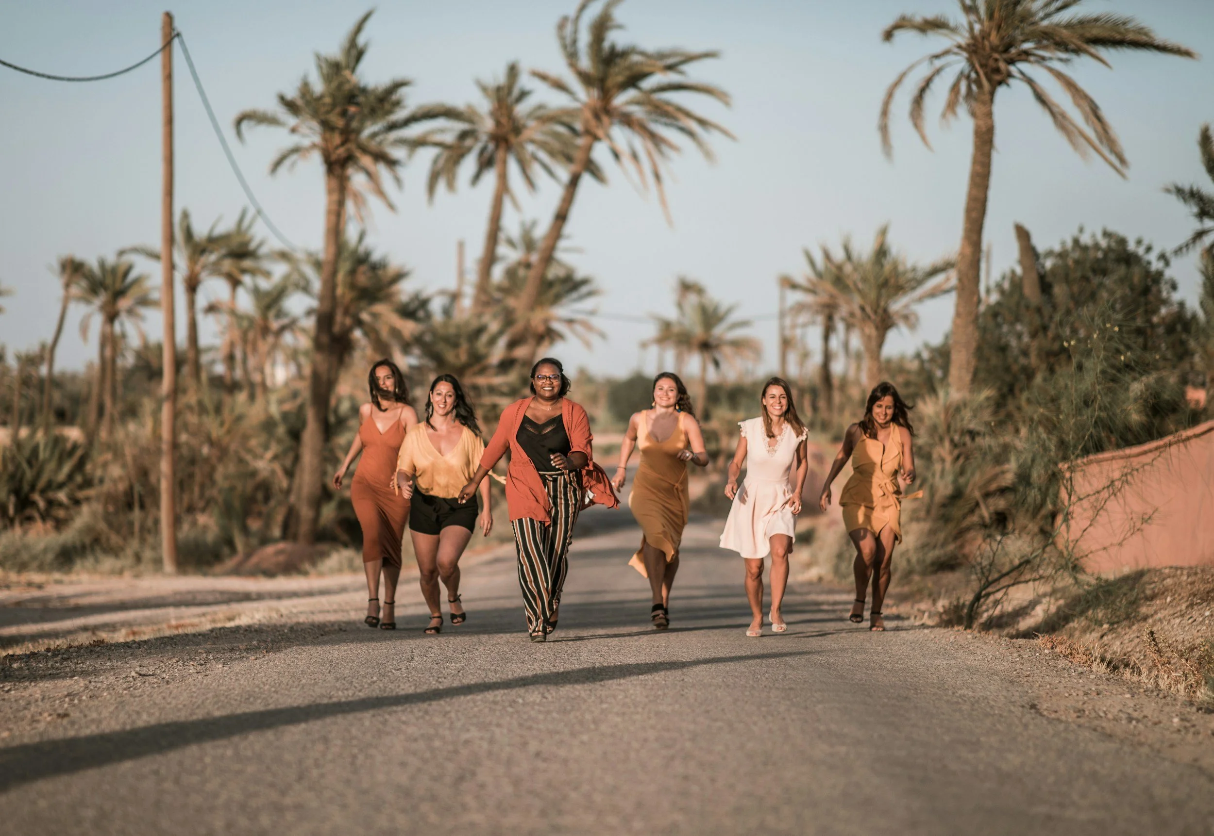Group of six joyful  multicultural women walking gleefully on a road in a desert-like area with palm trees in the background during sunset.