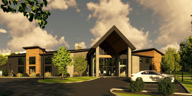 Modern building with large glass entrance and wooden accents, surrounded by trees and parking lot.