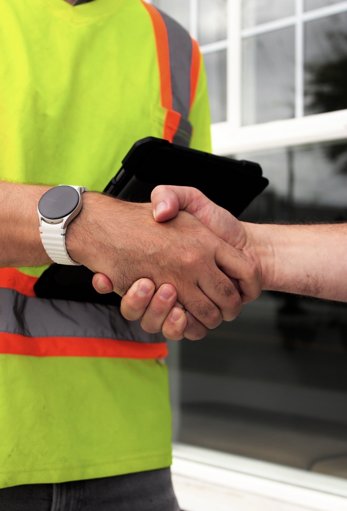 Two people shaking hands, one wearing a high-visibility safety vest, a wristwatch, and holding a tablet in an outdoor setting. This symbolizes relationship building.
