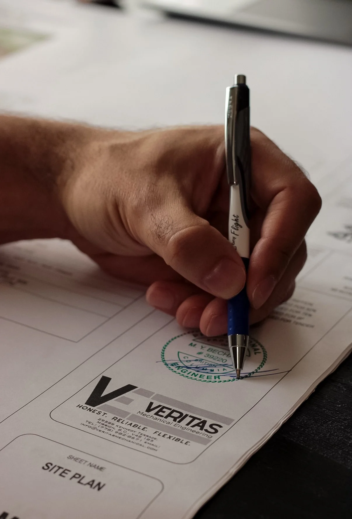 A person signing a document with a black and silver pen, the document has a green circular stamp with the word 'VERITAS' visible.