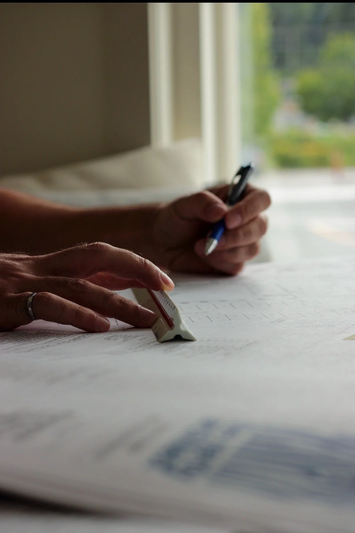 Close-up of two hands working on documents on a table near a window, with one hand holding a pen and the other using a highlighter.