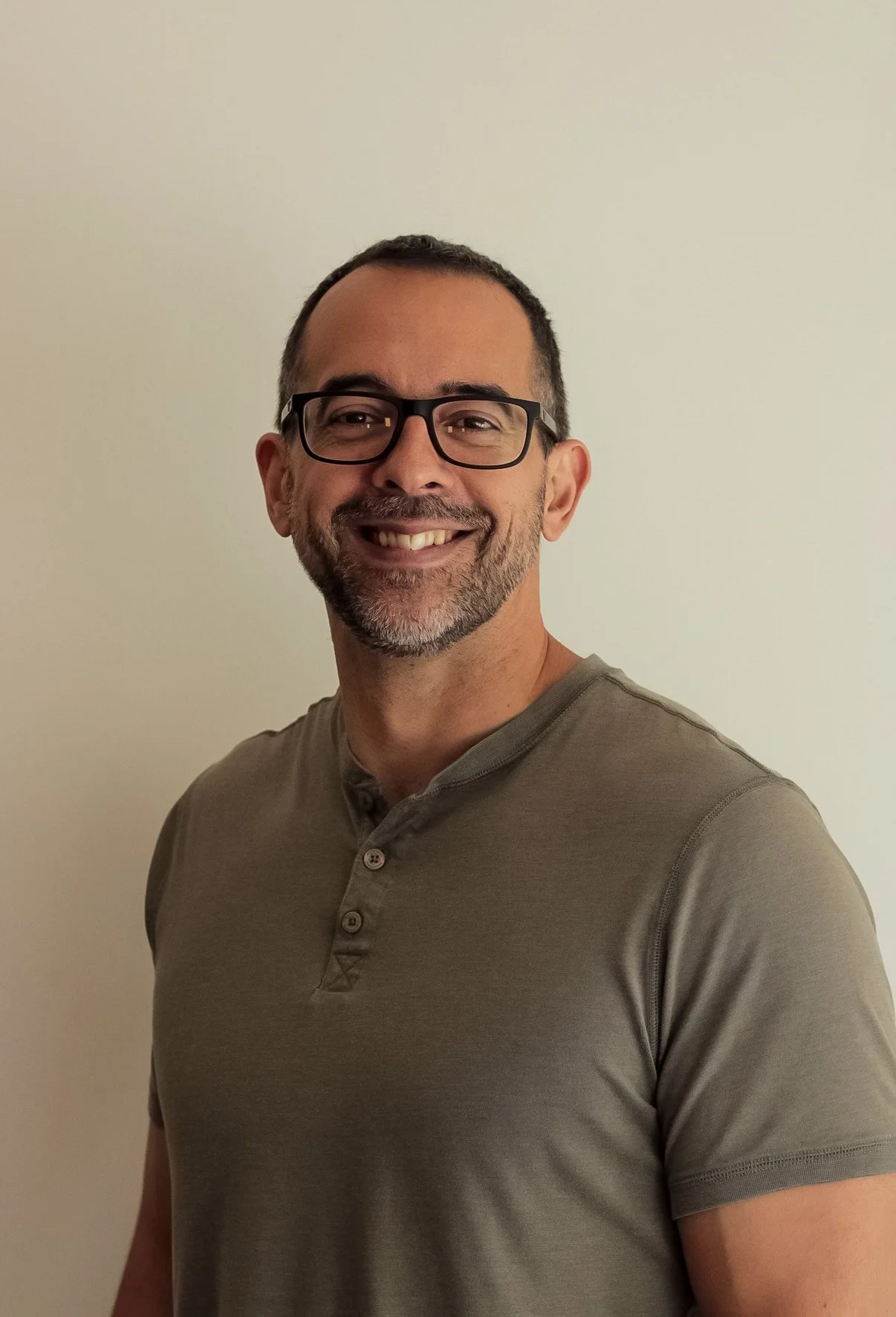 A middle-aged man with glasses, short dark hair, and a beard, smiling while wearing a short-sleeved grey shirt, standing against a plain off-white wall.