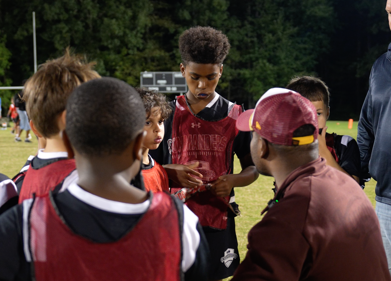 A youth football team in a huddle during a game at night, with coach talking to players on a field.