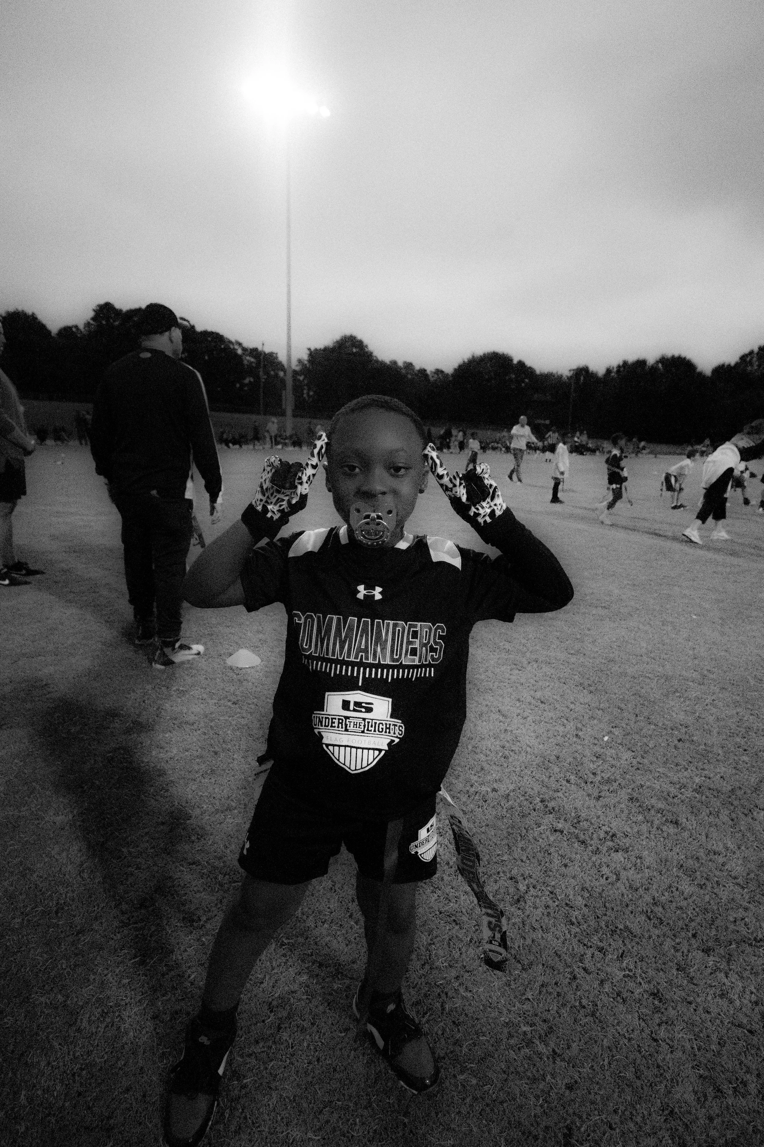 A young boy standing on a sports field at dusk, holding a football helmet under his arm, wearing a sports jersey with 'Commanders' written on it, and using a mouth guard.