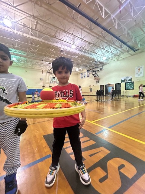 A young boy in a gymnasium holds a yellow tennis racket with a red ball on it, wearing a red shirt and sneakers.