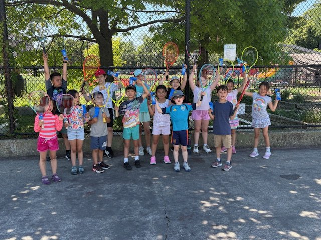 Group of children holding tennis rackets and water bottles, standing outdoors on a tennis court with a fence and trees in the background.