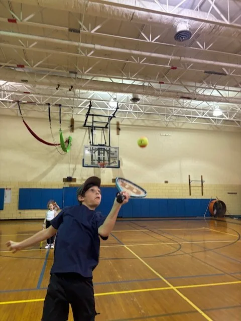 A boy playing pickleball in a gymnasium, swinging a paddle towards a ball.