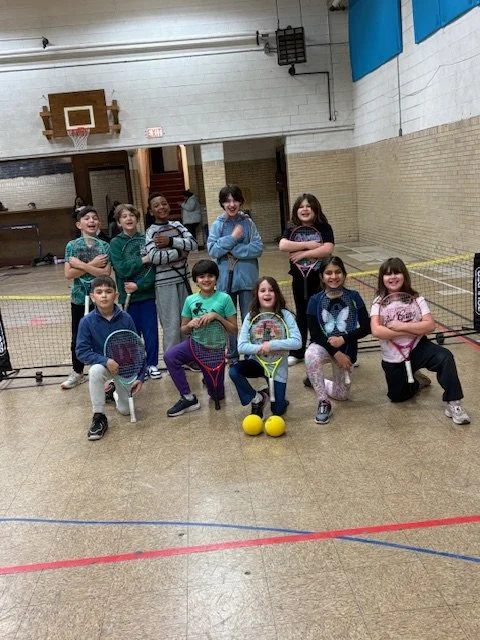 Group of children in a gymnasium, holding tennis rackets and tennis balls, posing for a photo.
