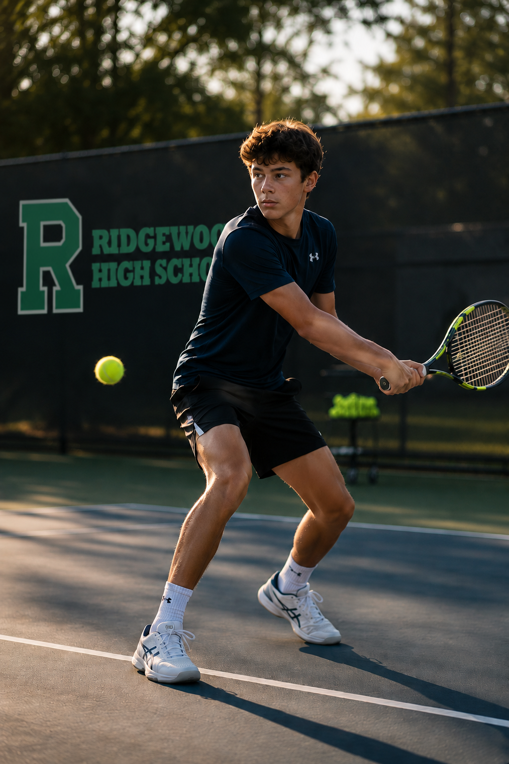 A young male tennis player in black athletic clothing and white shoes preparing to hit a tennis ball on an outdoor court with a Ridgewood High School sign in the background.