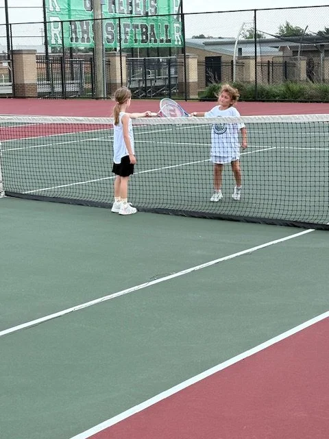Two young girls are playing tennis on an outdoor court, with one girl inside the court and the other girl standing outside near the net, holding a tennis racket.