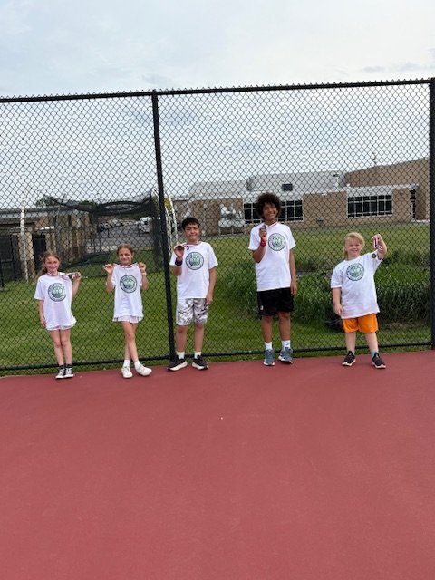 Five children standing in a row on an outdoor basketball court, holding up hand signs, with a chain-link fence and school building in the background.