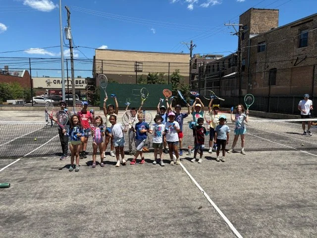 Group of children on an outdoor tennis court holding tennis rackets.