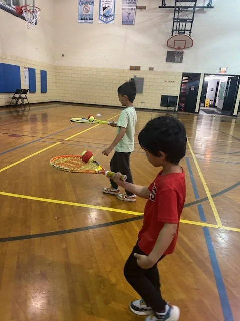 Two young boys playing badminton with rackets and shuttlecocks in an indoor gymnasium with basketball hoops and banners hanging from the ceiling.