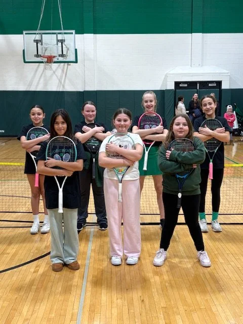 Group of seven young girls standing on an indoor tennis court, holding tennis rackets, with a basketball hoop and green and white walls in the background.