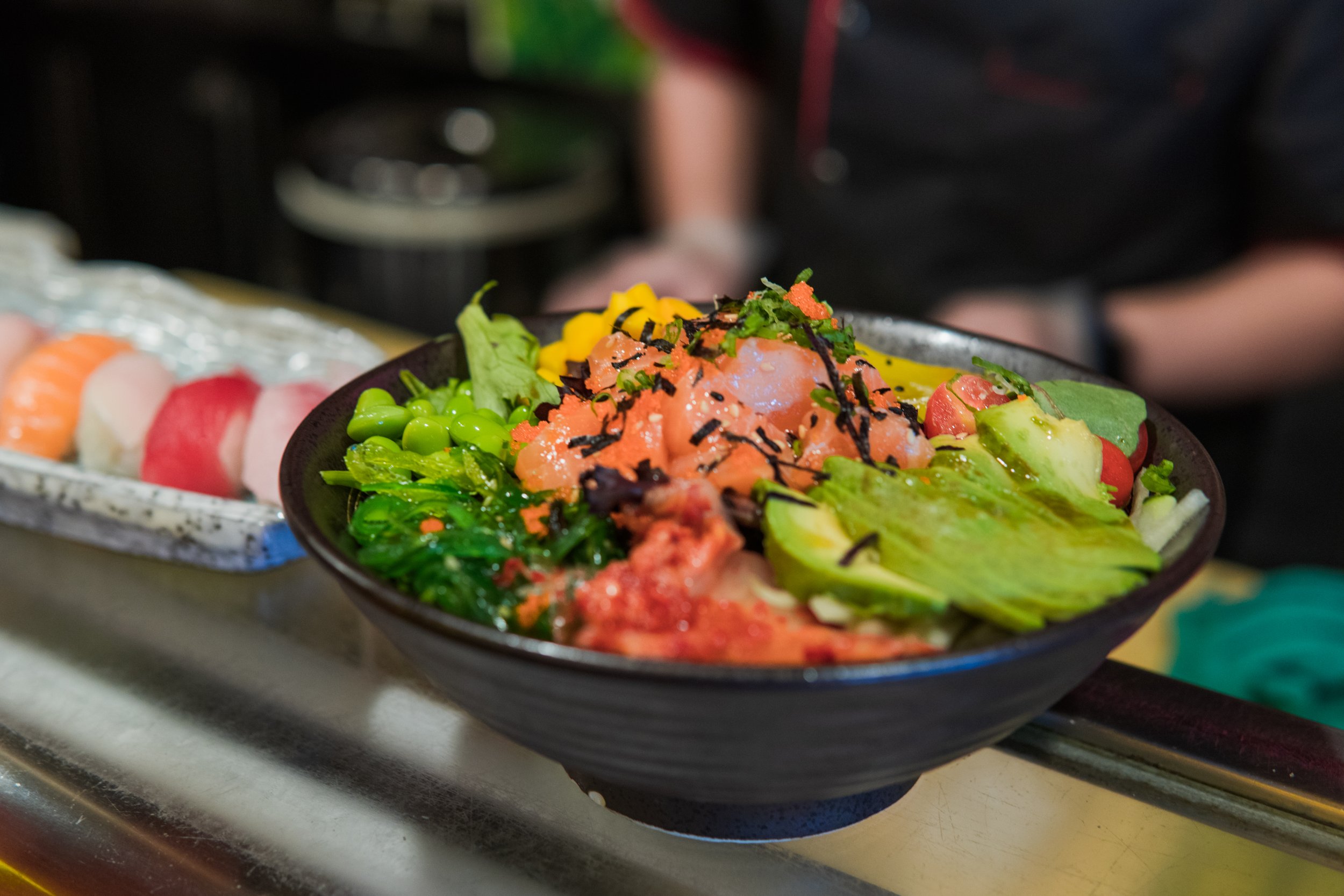 A black bowl filled with various fresh ingredients including lettuce, bright orange fish roe, shredded nori, chopped green onions, diced tomatoes, and other vegetables, placed on a kitchen counter.