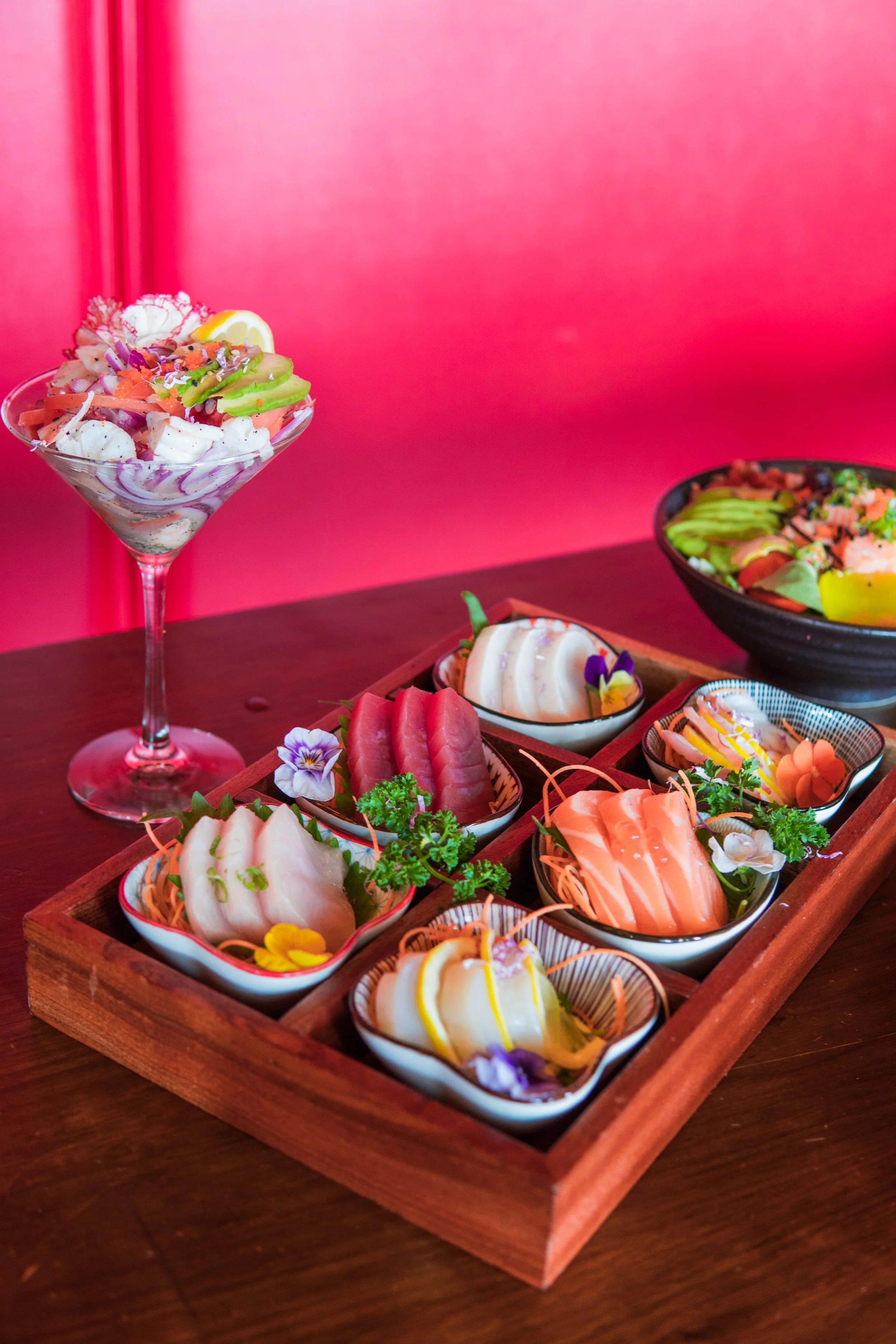 Assorted sushi and sashimi dishes served in small bowls and on a wooden tray, with vegetables, garnishes, and decorations on a dark wooden table against a bright pink background.