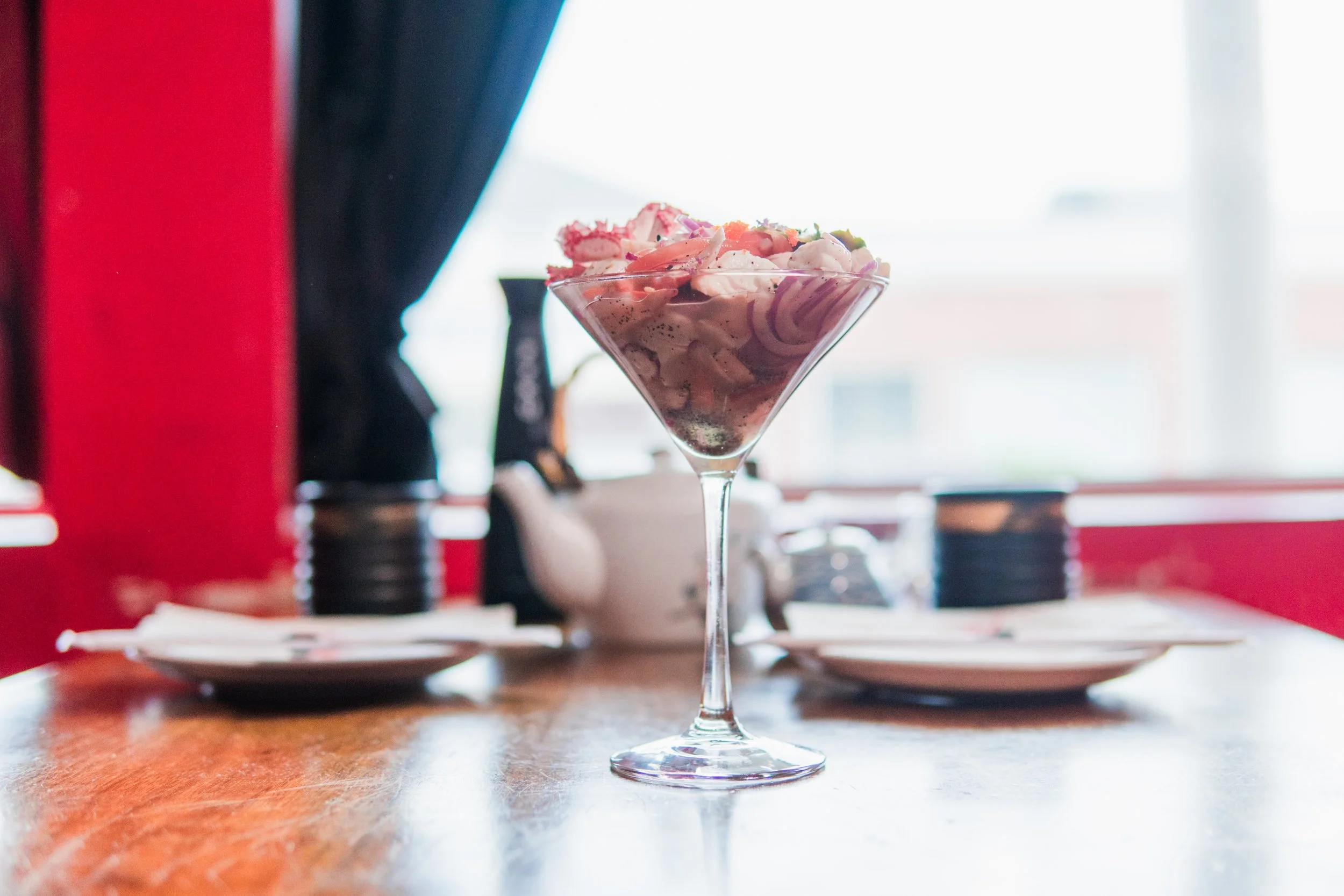 A martini glass filled with a pink cocktail or dessert topped with pink and white shavings, possibly candy or ice cream, placed on a wooden table inside a restaurant with blurred background.
