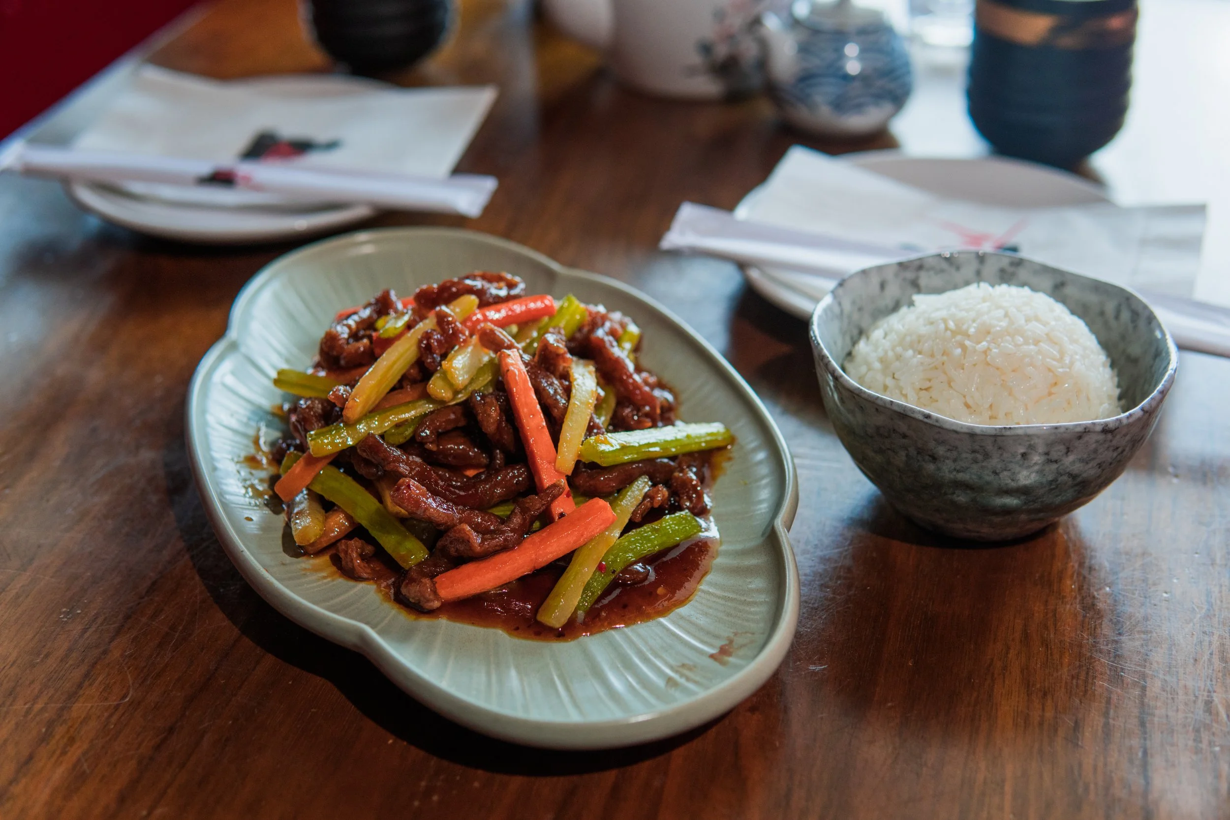 A plate of stir-fried beef with colorful vegetables and a bowl of white rice on a wooden table.