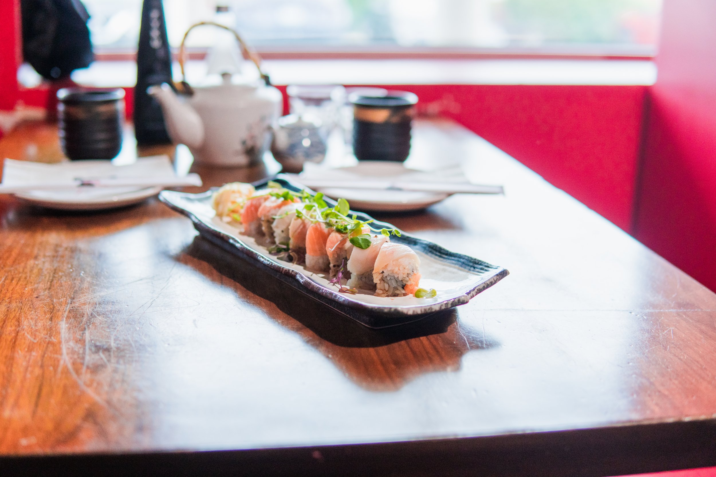 A restaurant table with a rectangular plate of sushi, including slices of fish and garnishes, on a wooden table in front of a red booth with a window in the background.