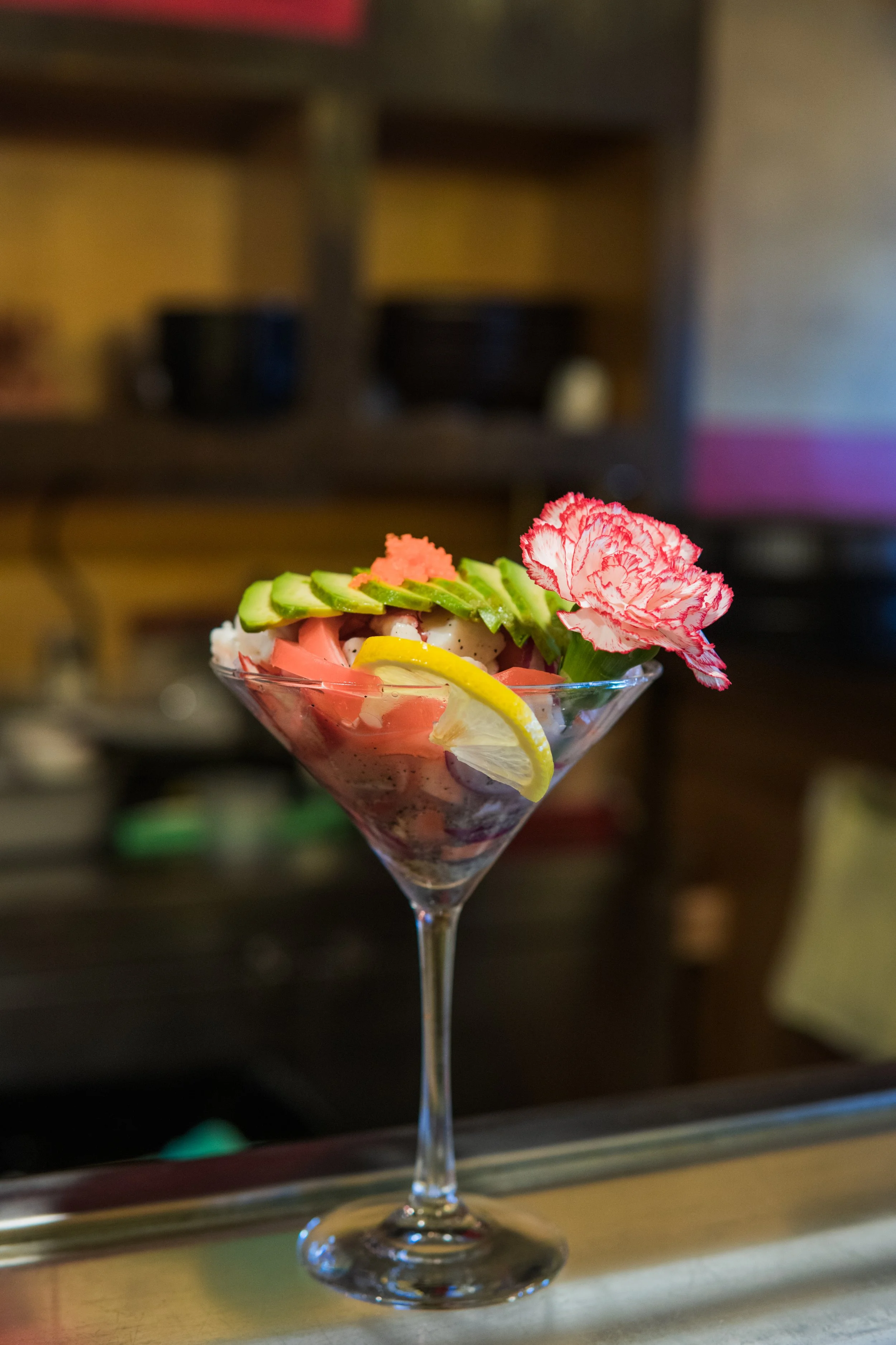A cocktail glass with a fruit garnish, including cucumber slices, lemon slices, pickled ginger, and a pink carnation flower, on a bar counter.