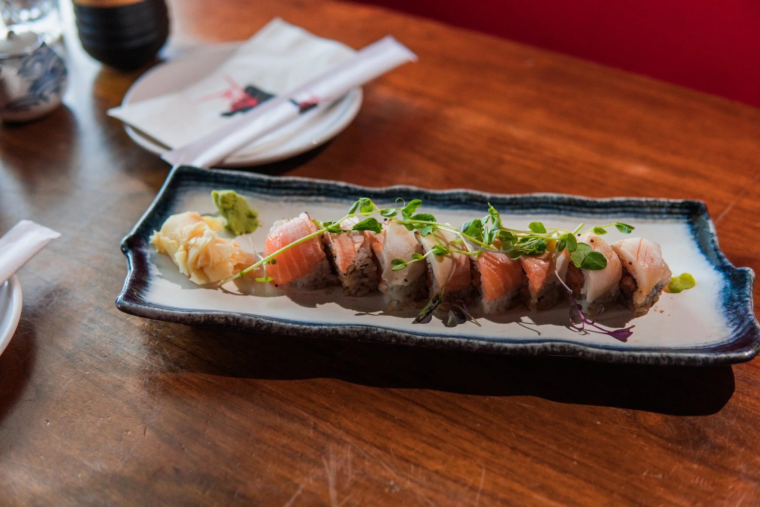 A plate of sushi rolls garnished with microgreens and accompanied by wasabi and pickled ginger, on a dark rectangular tray on a wooden table.