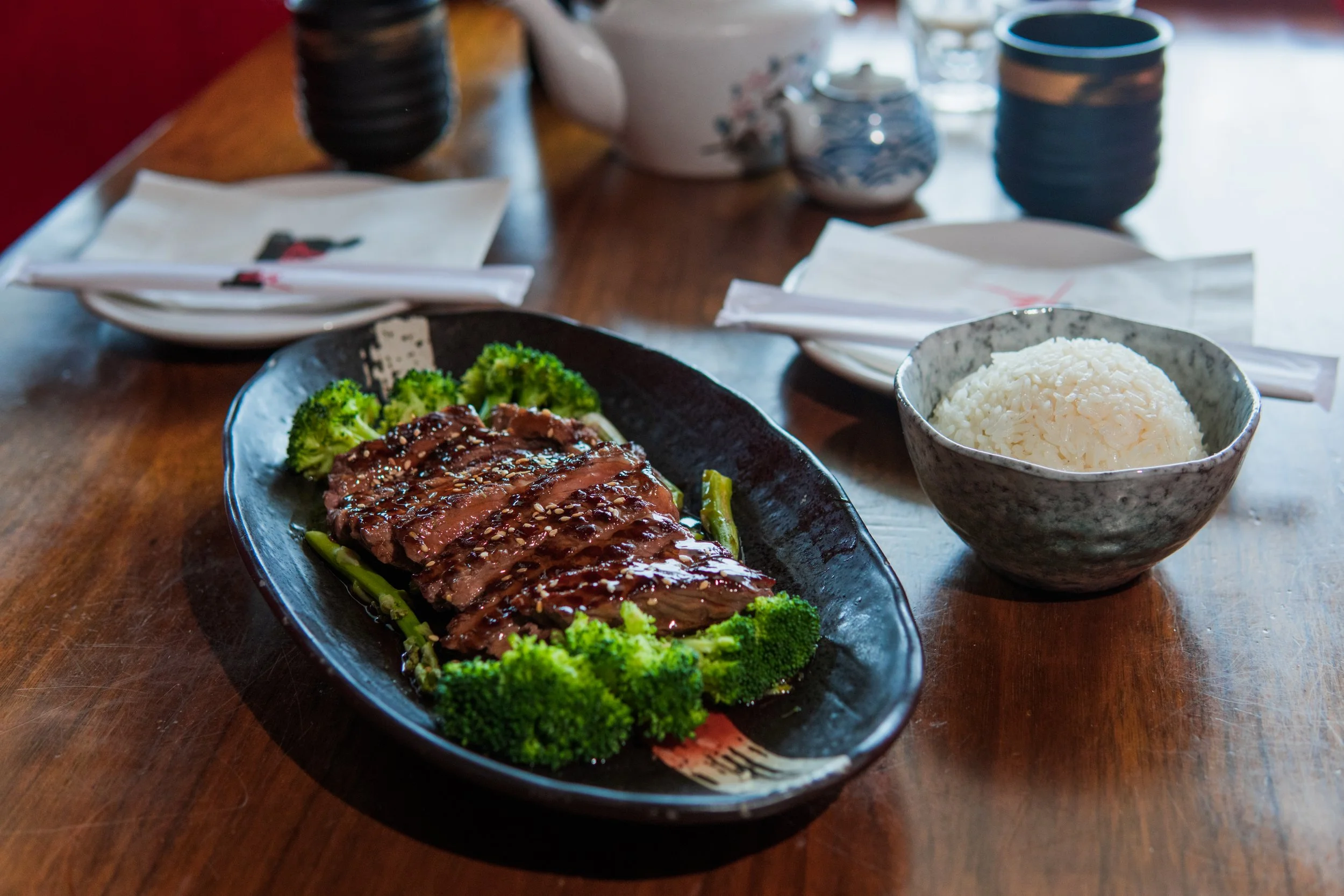 A plate of glazed beef with sesame seeds served over broccoli and green beans, white rice in a bowl, and various bowls and utensils on a wooden table.