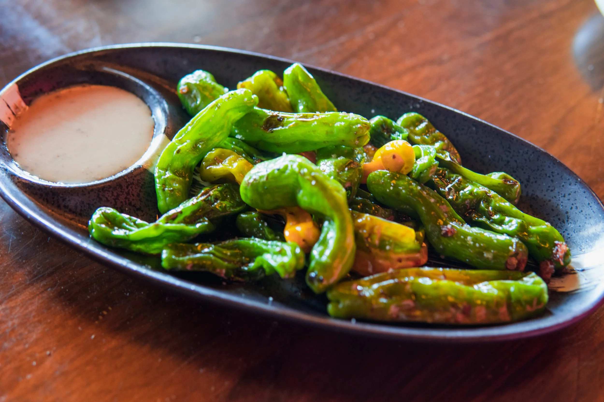 A black oval dish with a small container of creamy sauce and a serving of roasted green peppers on a wooden table.