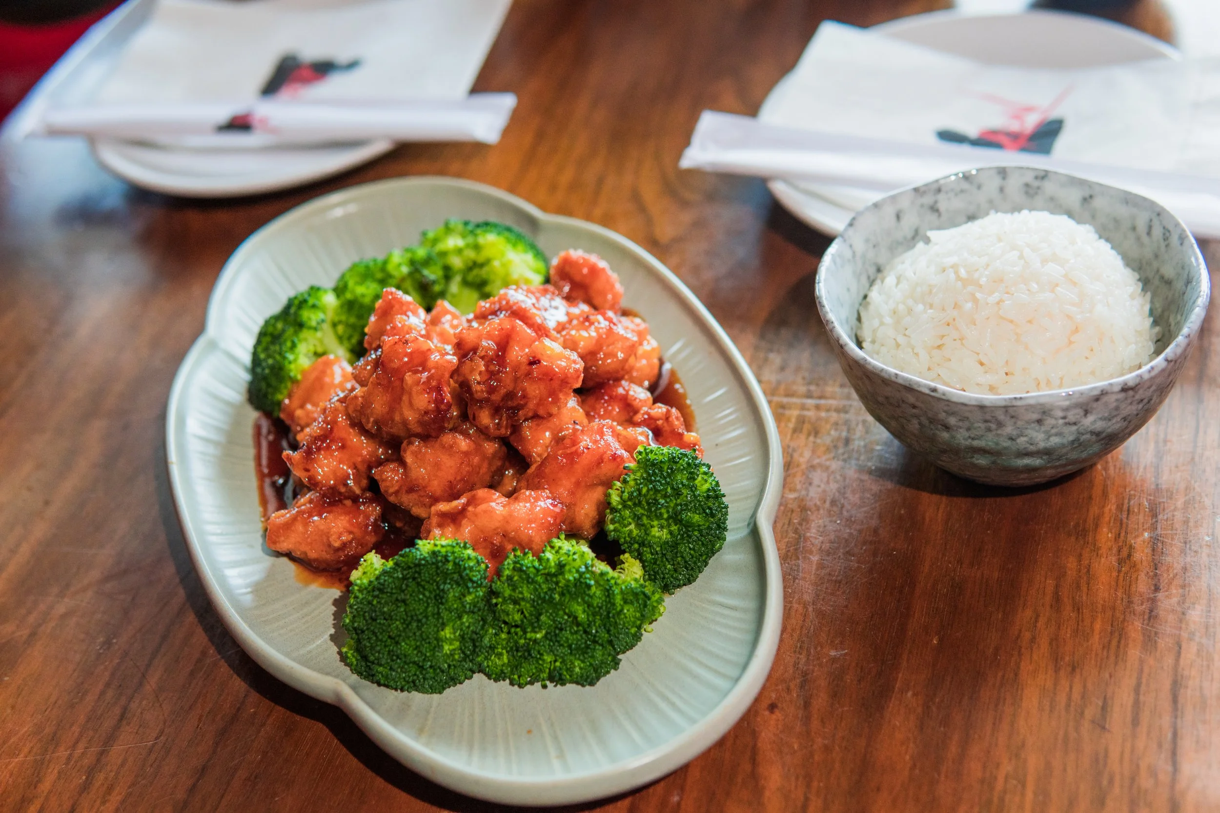 A plate of orange chicken with broccoli, a bowl of white rice, and chopsticks on napkins on a wooden table.