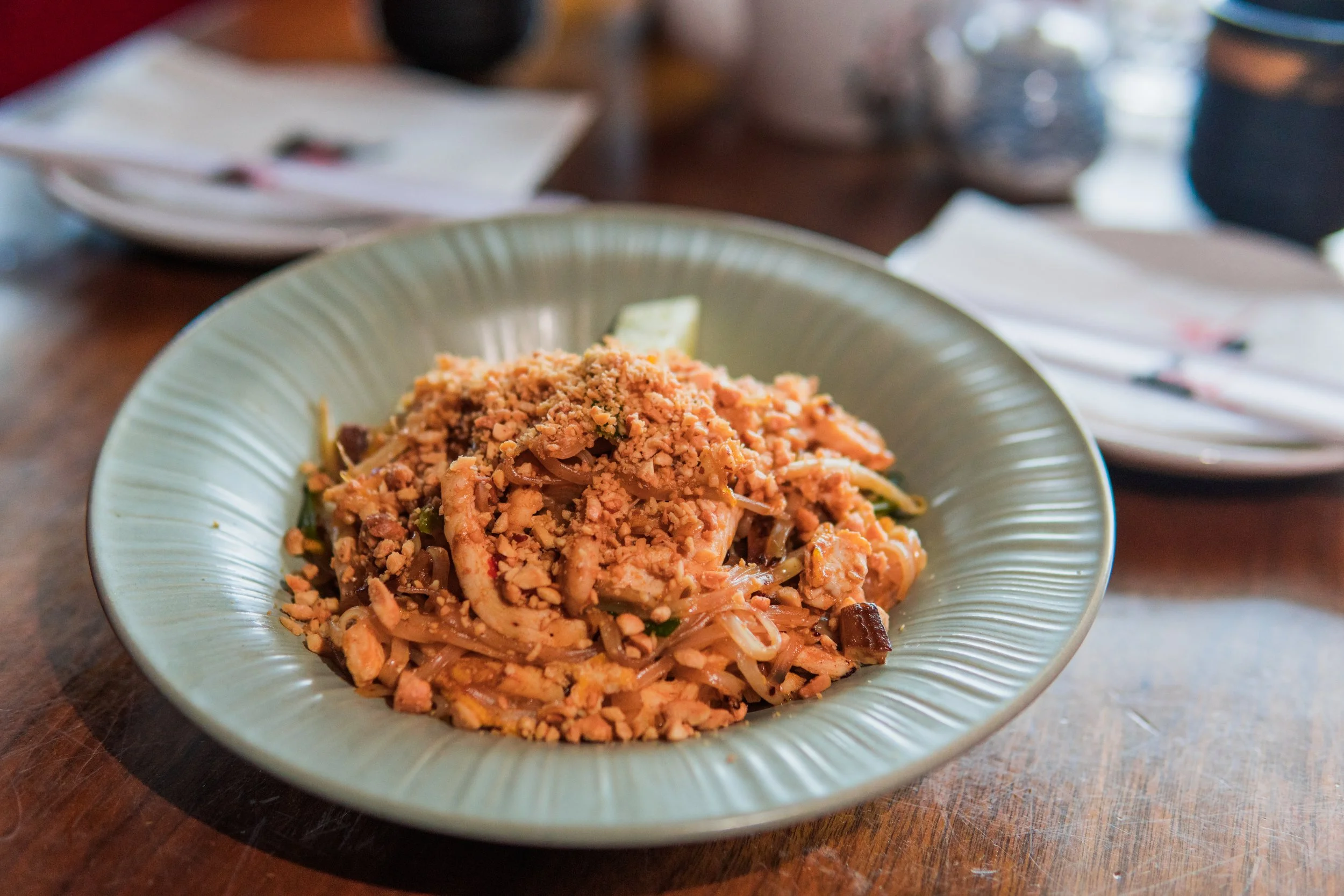 A bowl of Asian noodle dish topped with crushed peanuts, served on a table with napkins and utensils in the background.