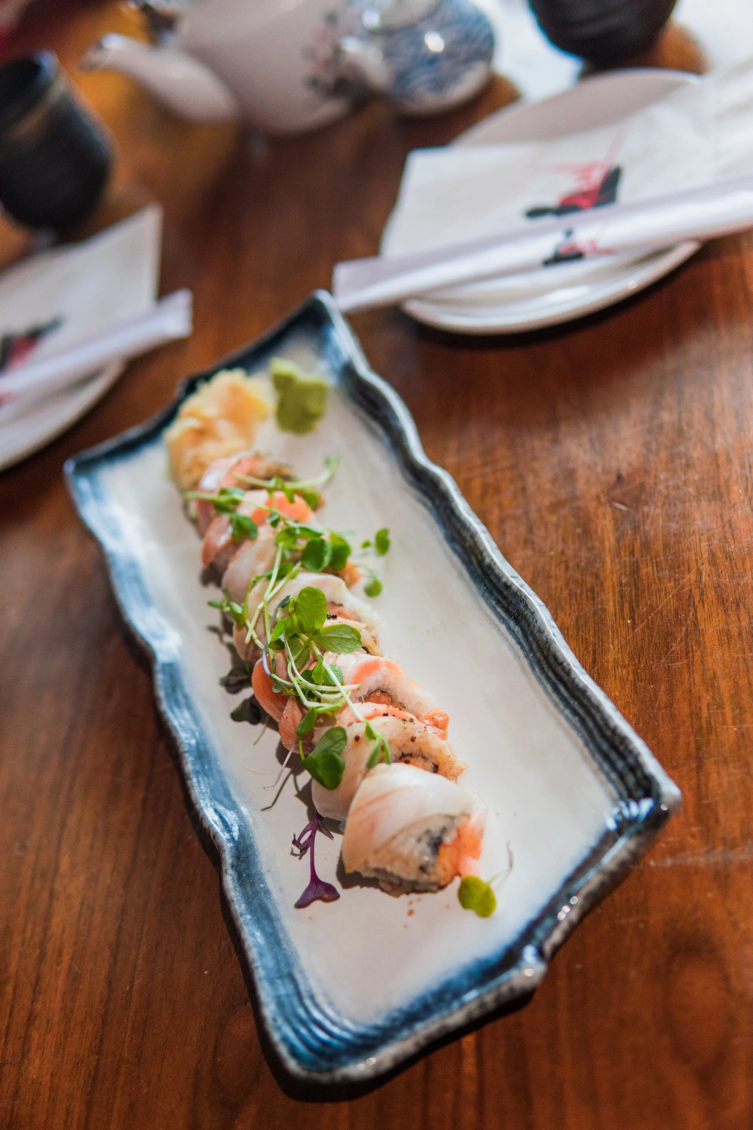 Sushi roll pieces garnished with microgreens served on a rectangular ceramic plate placed on a wooden table, with a small dish of wasabi and ginger nearby.