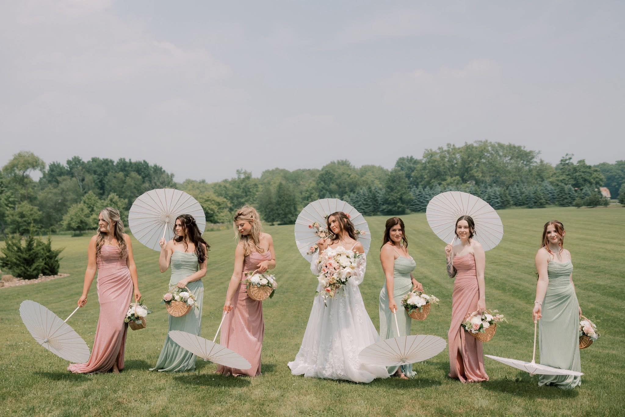 A group of women dressed in pastel-colored dresses holding floral bouquets and white parasols, standing outdoors on a grassy field with trees in the background.