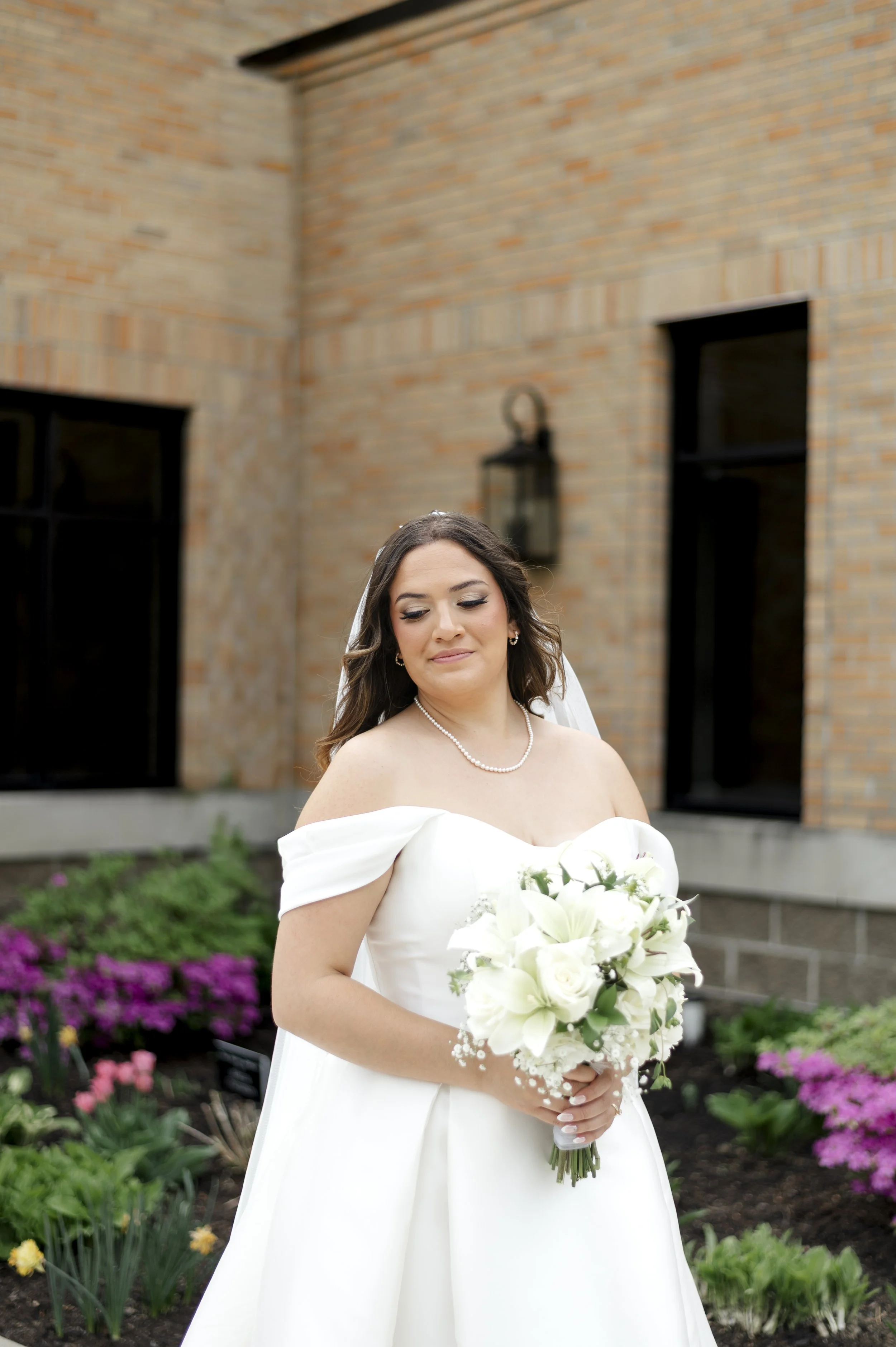 A bride in a white wedding dress holding a bouquet of white flowers, standing outdoors in a garden with pink and purple flowers and a brick building in the background.